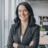 A woman with shoulder-length black hair wearing a smart blazer smiling at the camera in a modern office.